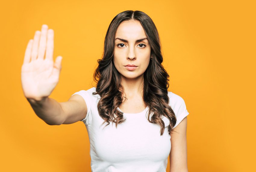 brunette woman looking serious holding hand out in front of her in a stop gesture - yellow-orange background - setting boundaries